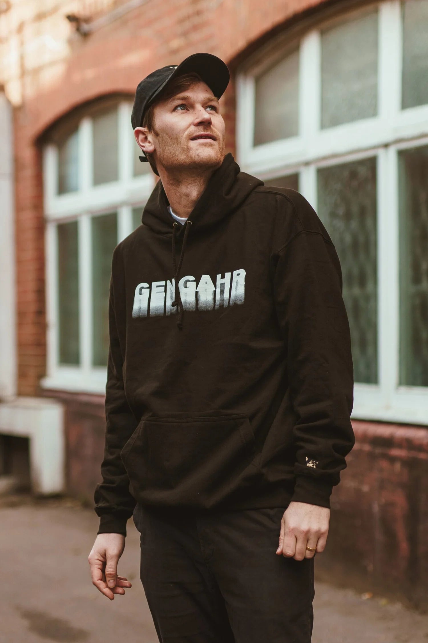Man wearing a black hoodie with 'GENGAHR' text, standing in front of a building with large windows.